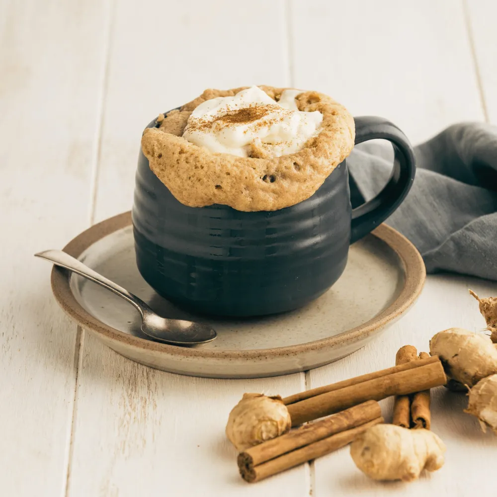 A completed Vanilla Chai Spiced Mug Cake recipe served warm in under two minutes. The mug cake is topped with whipped cream and ground spice, sitting next to cinnamon sticks on a rustic white table.
