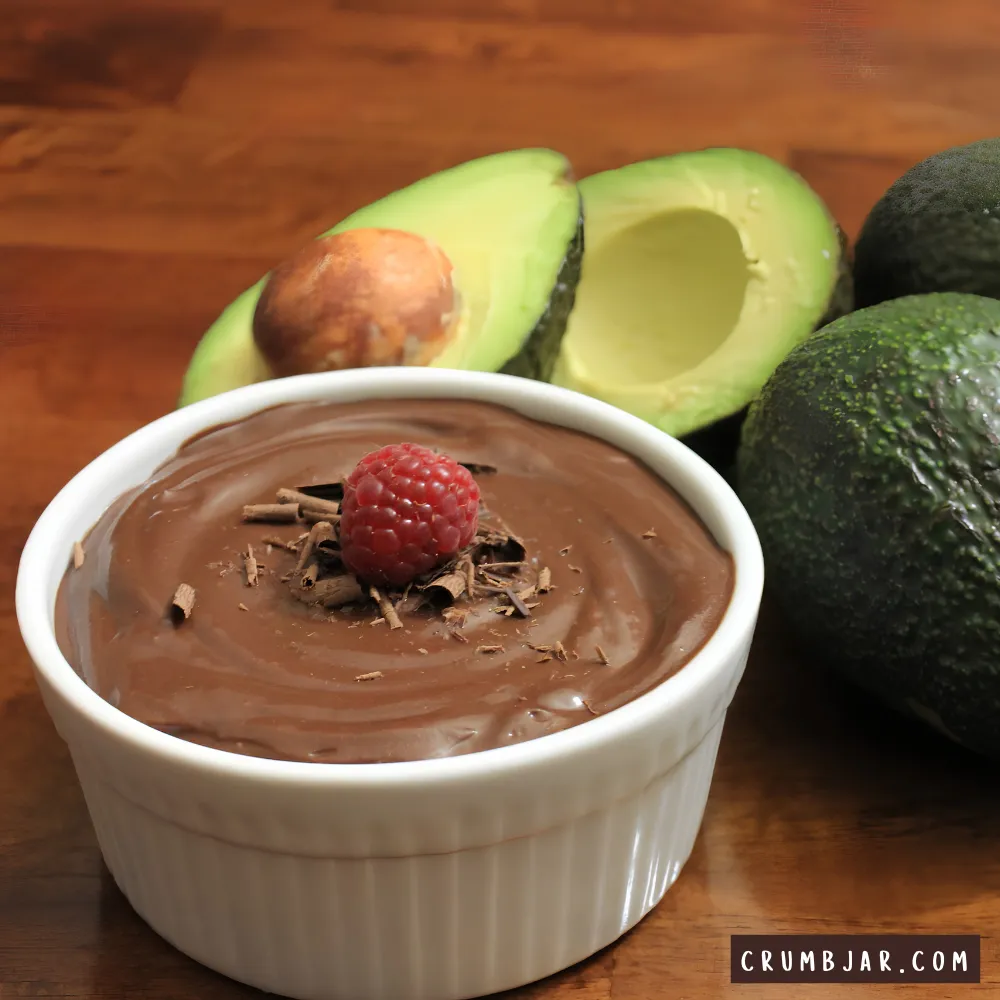 A close-up photograph of a smooth, glossy chocolate avocado pudding served in a white ribbed ramekin. The rich chocolate cream is topped with a single fresh raspberry and a sprinkle of dark chocolate shavings. In the background, fresh whole and halved green avocados rest on a warm wooden surface, showcasing the dessert's main ingredient.