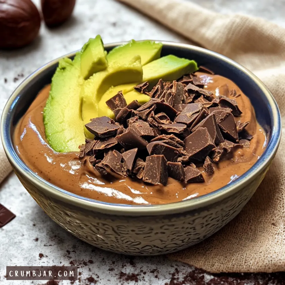 A close-up photograph of a thick, glossy chocolate avocado smoothie bowl served in an ornate, textured ceramic bowl. The creamy chocolate pudding is beautifully arranged with a fan of fresh green avocado slices on one side and a generous mound of roughly chopped dark chocolate chunks down the center. The bowl rests on a light grey textured surface alongside a soft linen cloth.