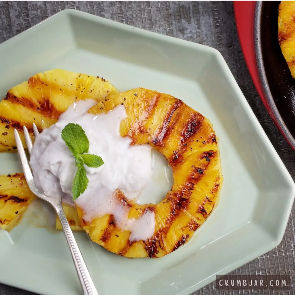 Warm grilled pineapple rings featuring charred grill marks served on a light green plate, topped with a dollop of fluffy white whipped coconut cream and a sprig of fresh mint, next to a silver dessert fork.