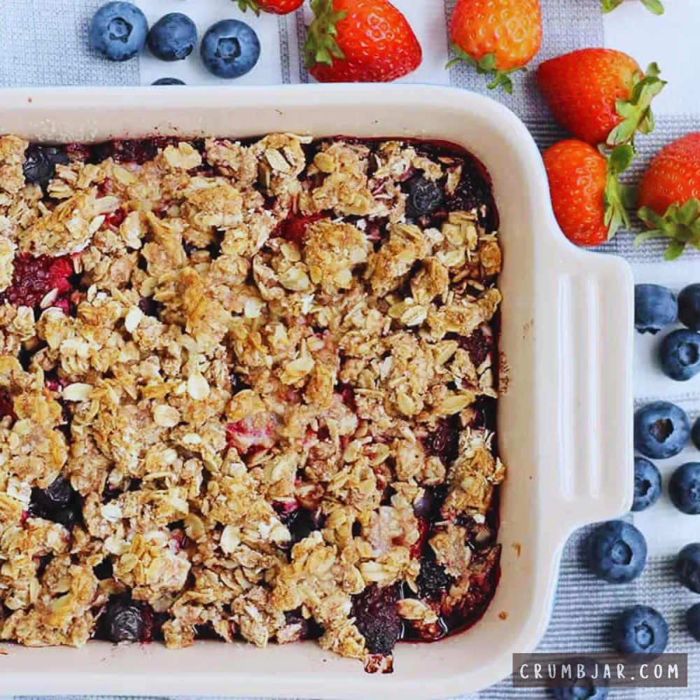 Freshly baked big-batch berry crisp topped with a golden oat streusel topping in a ceramic baking dish, surrounded by fresh strawberries and blueberries.