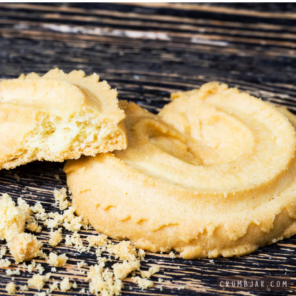 Close-up of a broken butter cookie showing a dry interior texture and loose crumbs on a dark wooden surface, illustrating "My cookies are dry and crumbly."