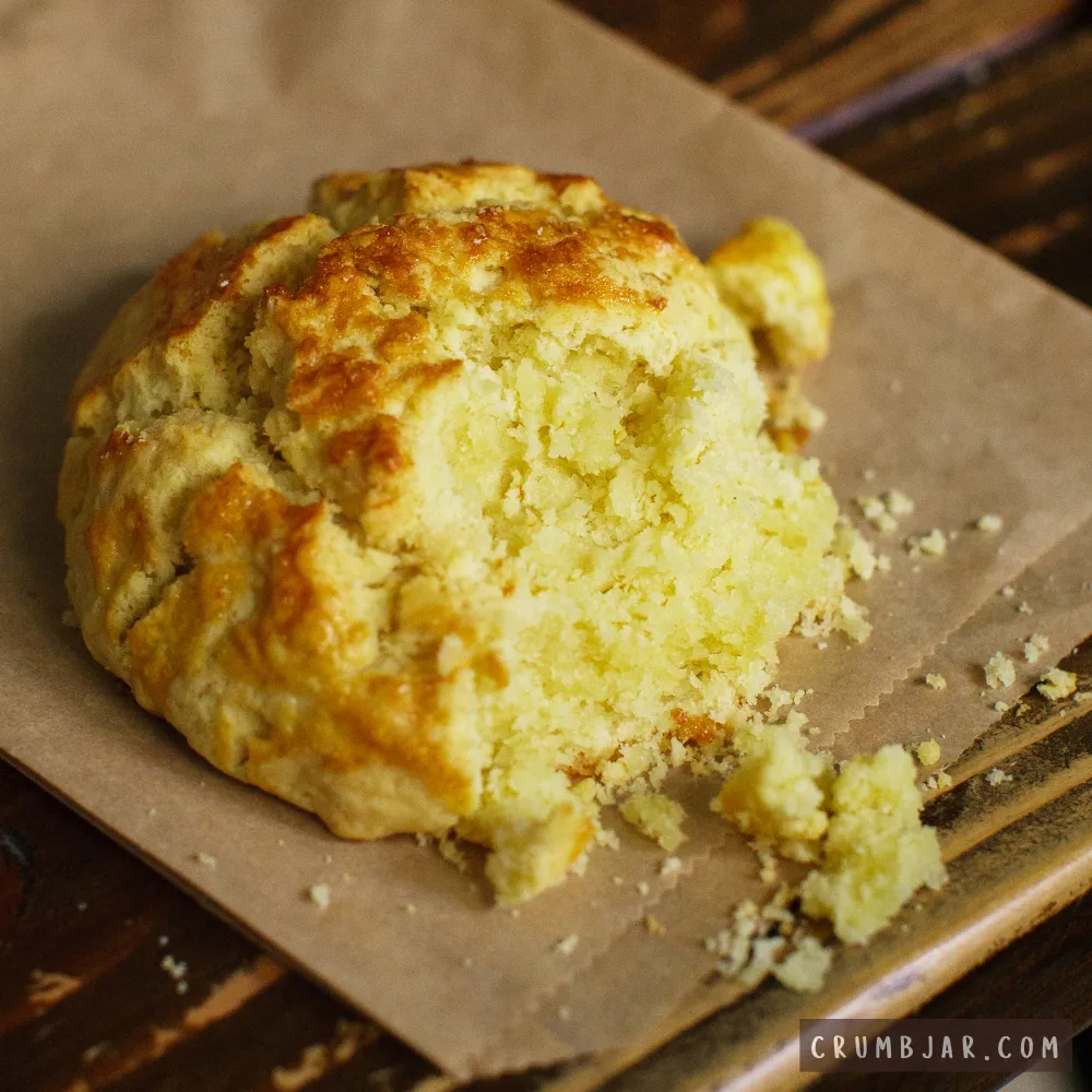 Close-up of a broken golden-yellow cake resting on brown parchment paper with loose crumbs falling away, illustrating "My cake is dry and crumbly."