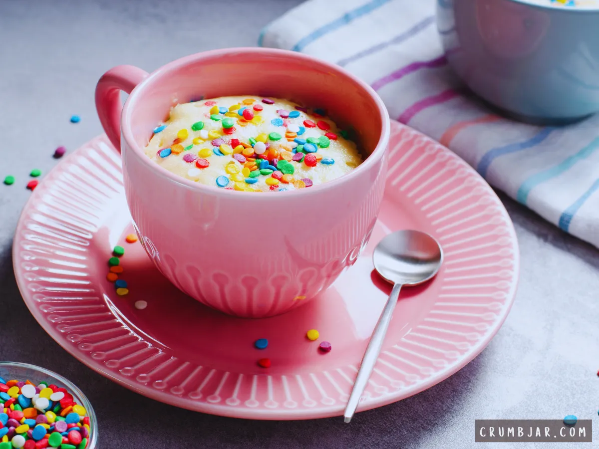A fluffy vanilla mug cake topped with colorful rainbow confetti sprinkles served in a pink ceramic mug on a matching pink saucer next to a small spoon, illustrating a Classic Vanilla Birthday Mug Cake.