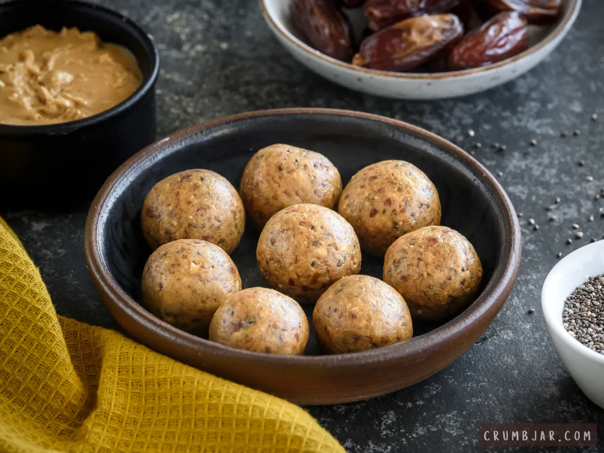 A dark ceramic bowl filled with round Chocolate Peanut Butter Energy Bites arranged on a dark counter alongside bowls of peanut butter, dates, and chia seeds.