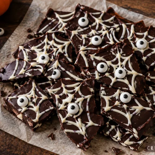 Homemade chocolate spiderweb bark with white chocolate web designs and candy eyeballs, arranged on parchment paper on a dark wooden background.