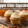 Three spiced apple cider cupcakes topped with swirls of cinnamon cream cheese frosting on a wooden cutting board, with fresh apples and a jug of apple cider in the background.