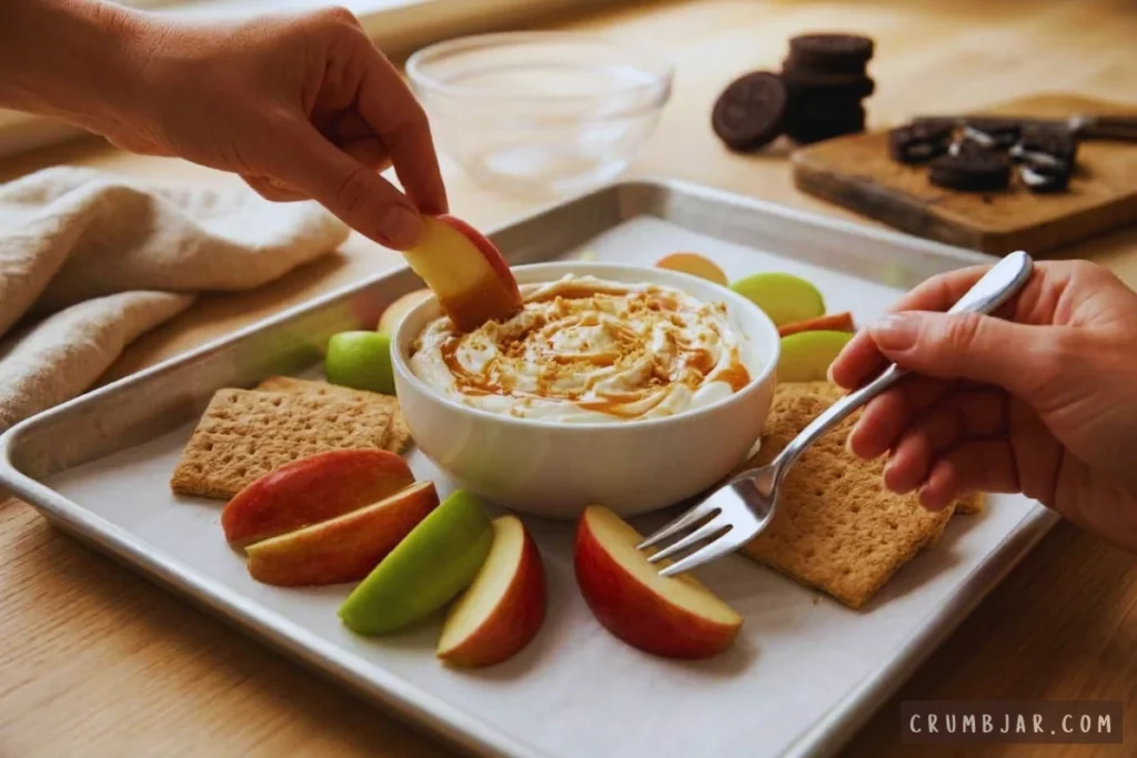 Dipping fresh red apple slices into a bowl of caramel cheesecake dip arranged on a serving tray with graham crackers.